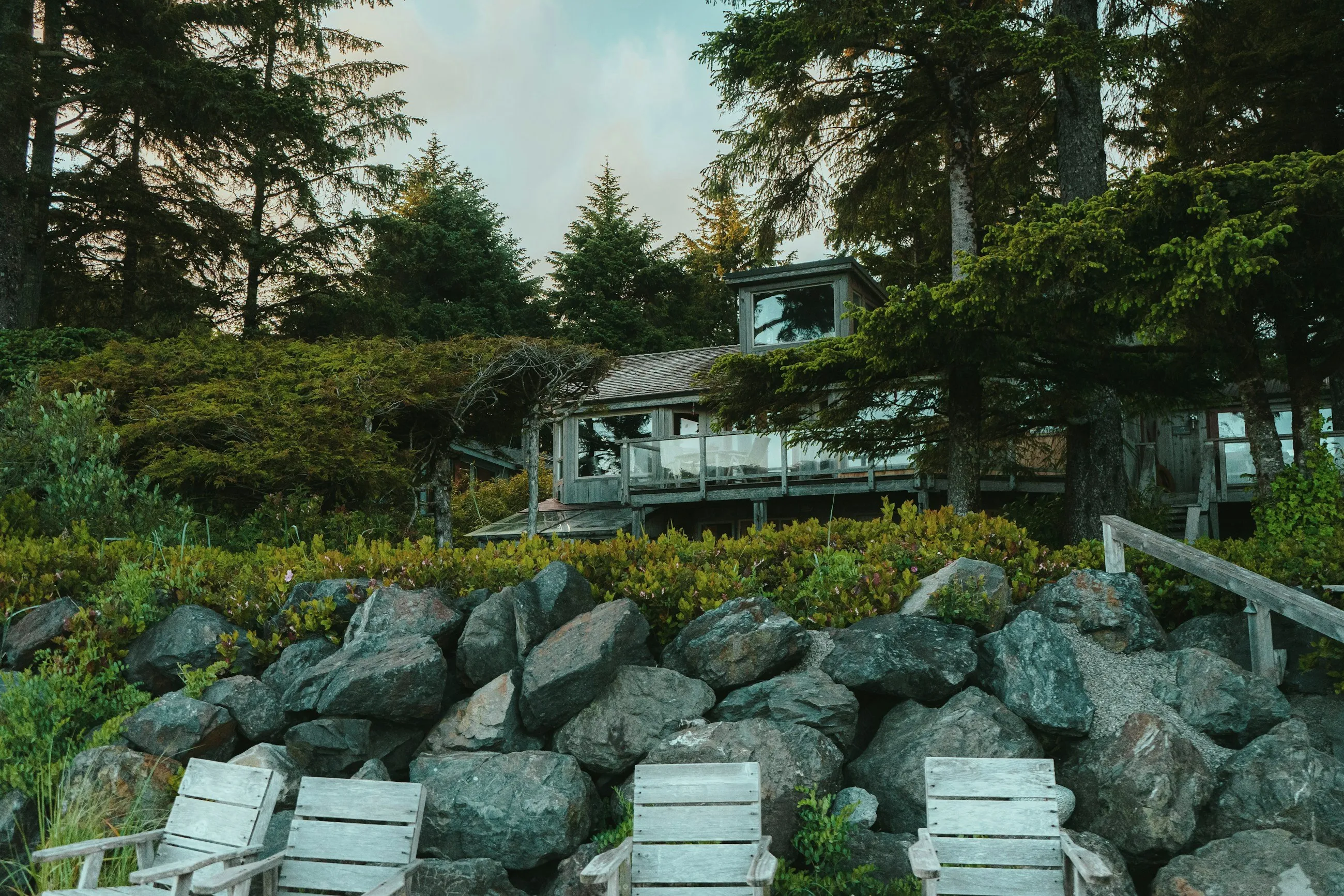 A cedar-and-glass house set back among Sitka spruce on the BC coast, its terrace held by a hand-placed granite wall with salal and native shrubs planted through it, weathered timber chairs on the gravel below.