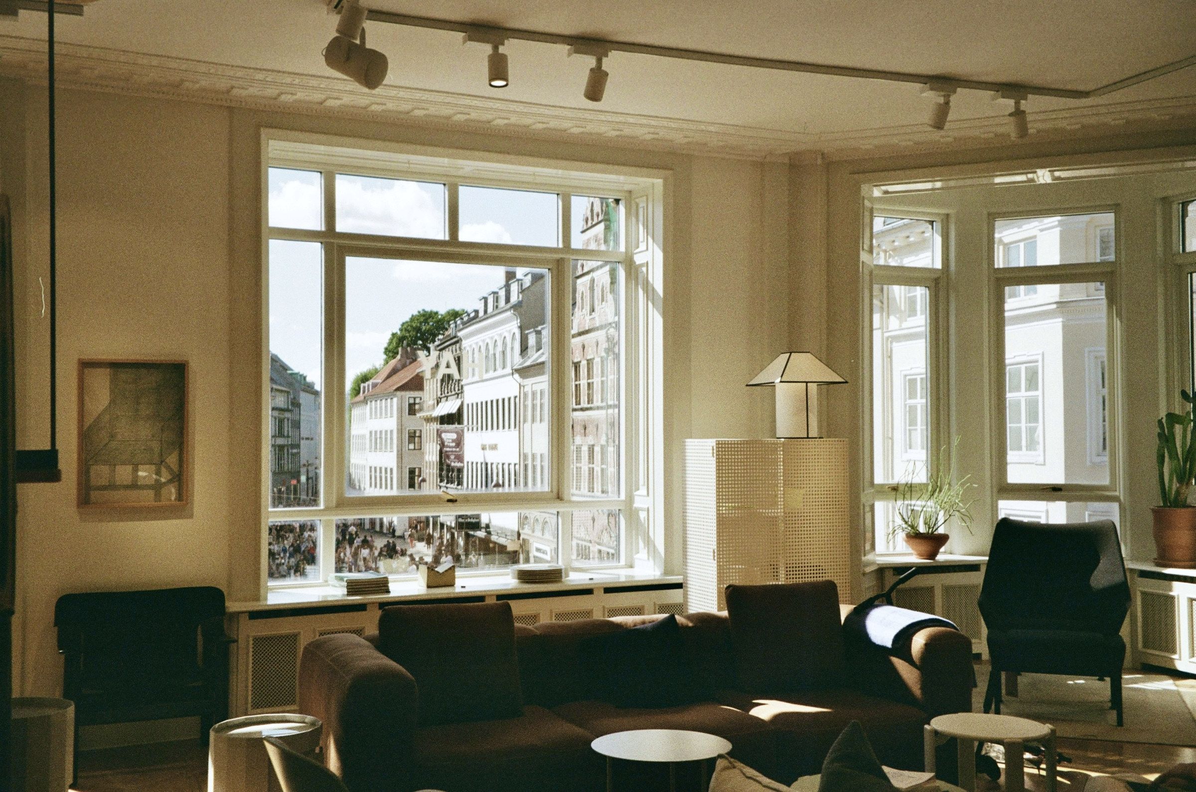 A Copenhagen apartment lounge in late afternoon sun: a long low sofa and two armchairs under tall white-framed sash windows, a paper-shade table lamp, plants on the sill, and the city rooftops beyond the glass.