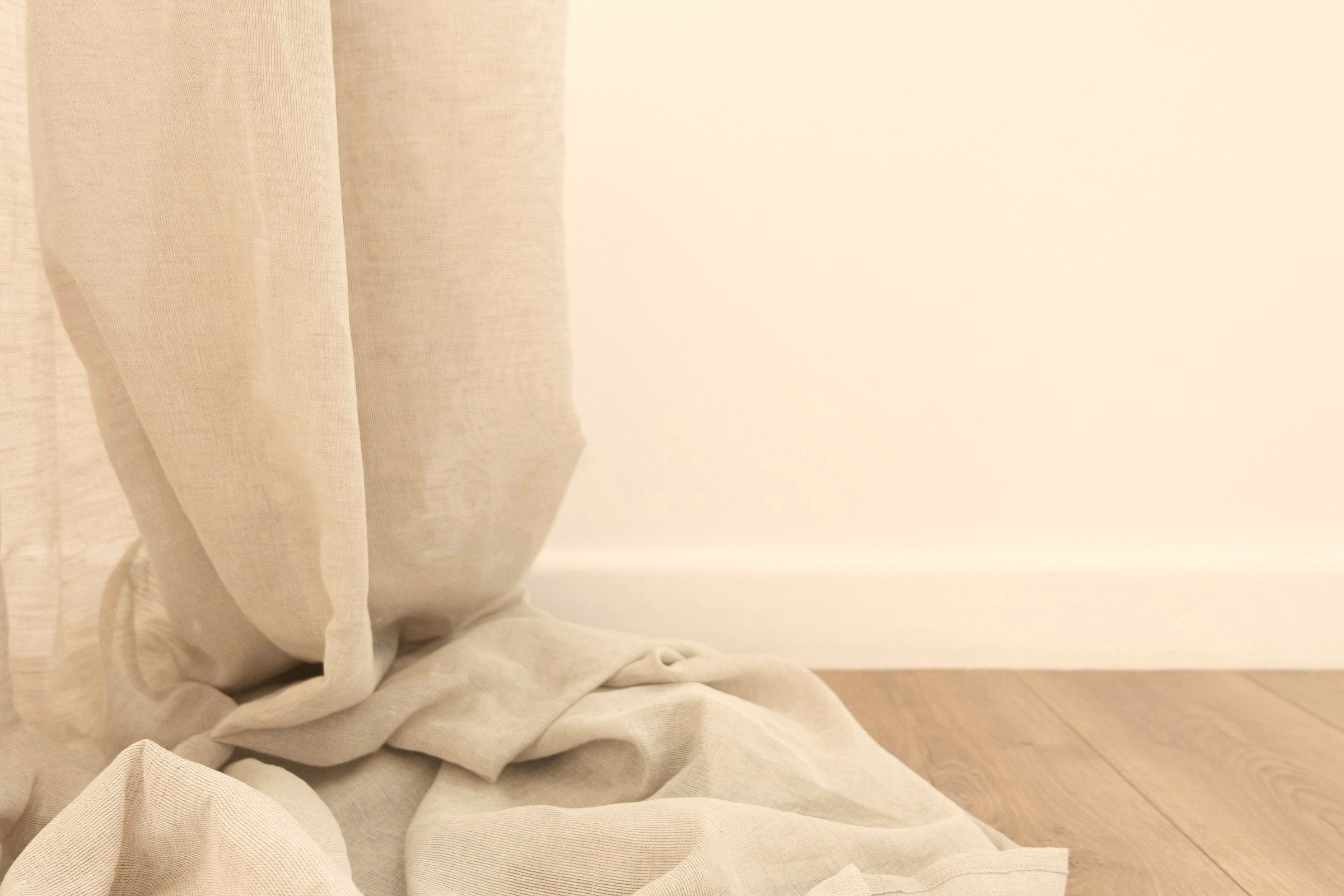 An undyed linen curtain falling in a deep fold and pooling on an oak floor.