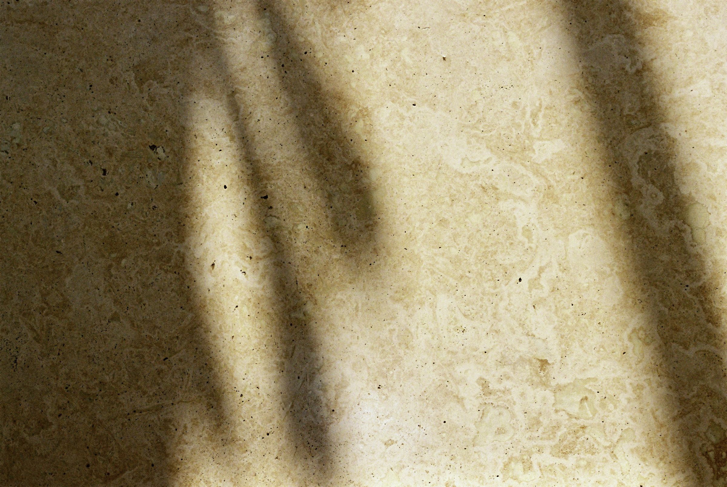 A mottled cream travertine surface, its open pores and bedding lines crossed by soft window shadows.