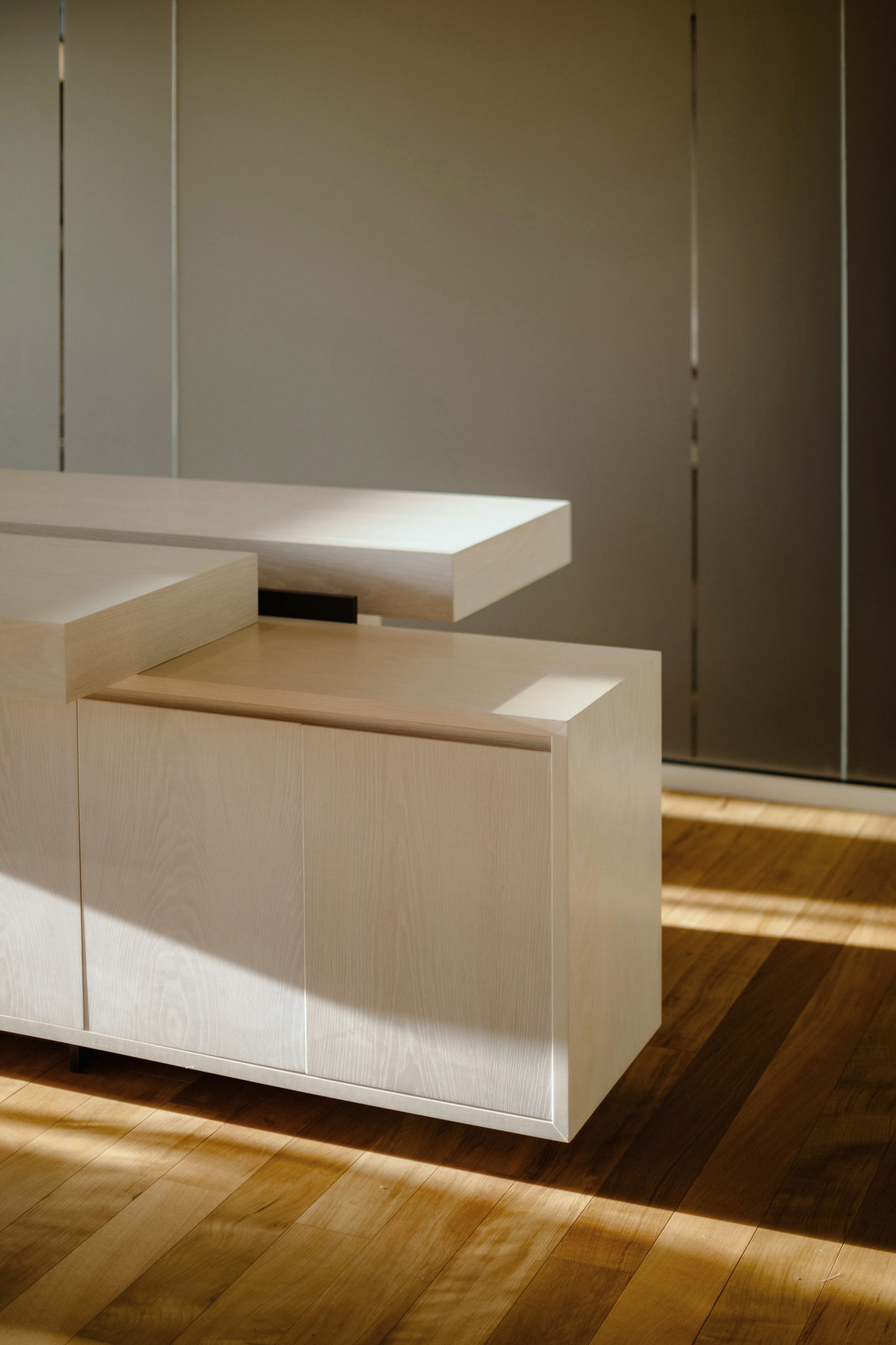 The lobby desk: a limed-oak counter carcase with its transaction shelf cantilevered over it, standing free on a plank floor in hard raking afternoon sun.
