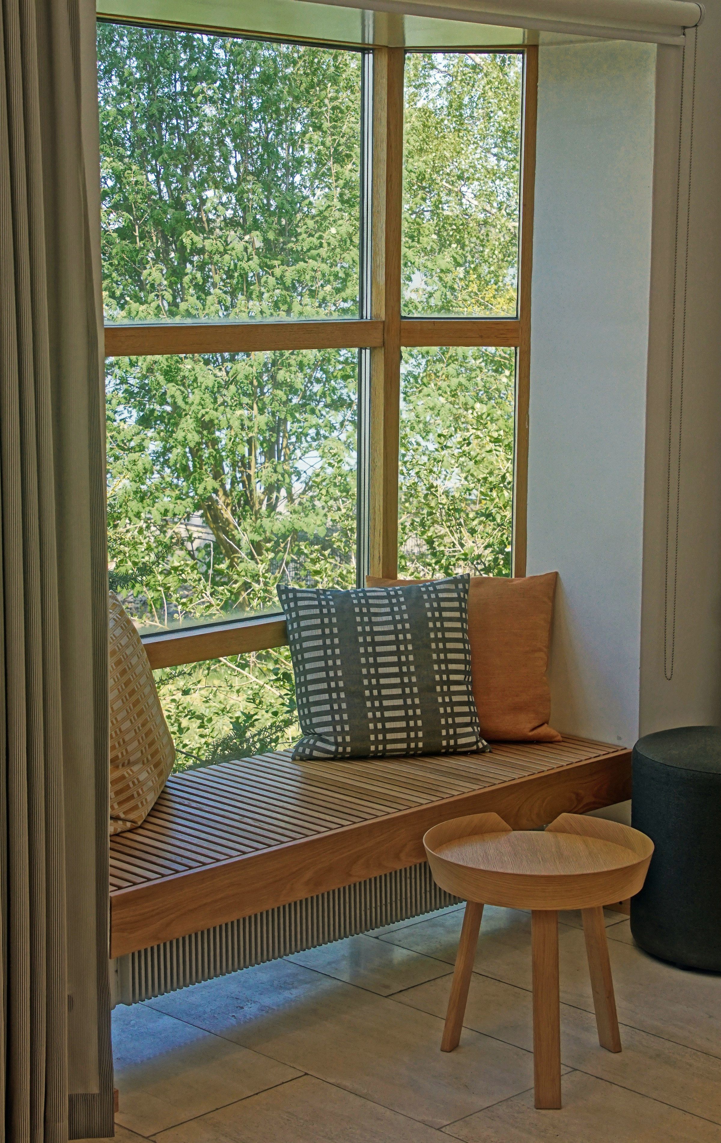 A window seat at Hotel Søgrund: a slatted oak bench built into the glazed corner, cushions in charcoal and rust against the linen curtain, an oak stool and a felted wool pouf on the travertine floor, birches filling the window.