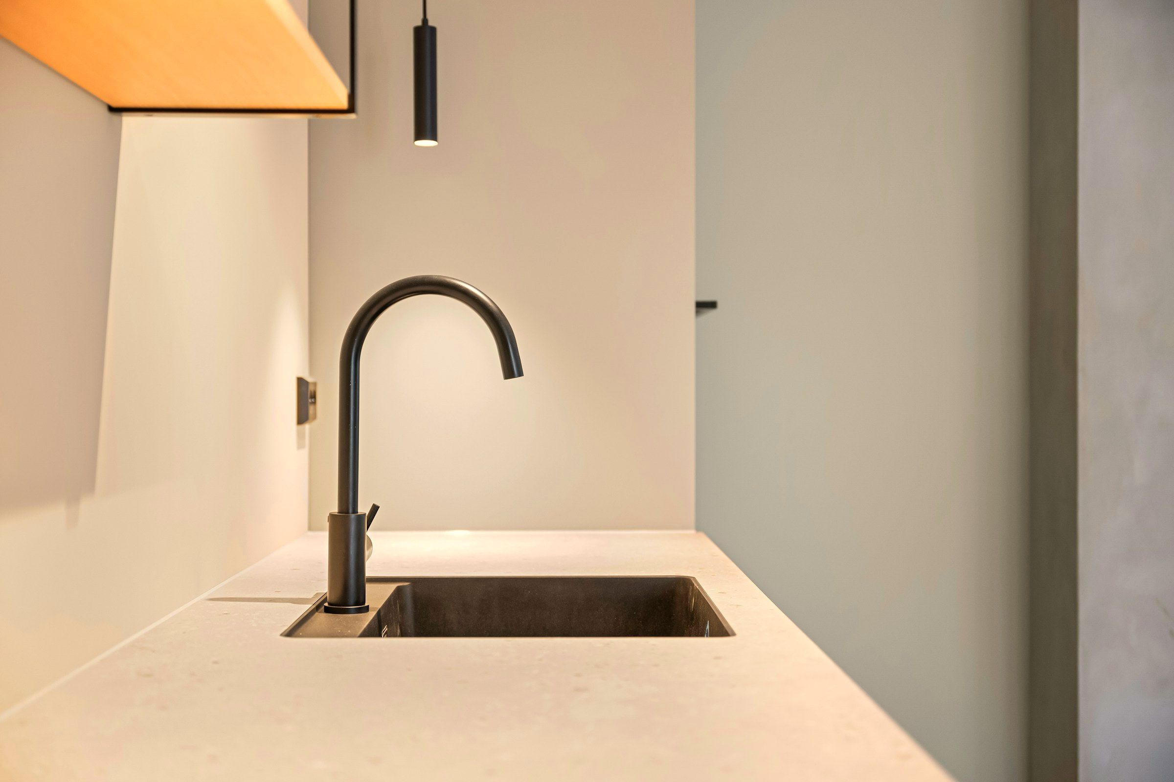 The kitchen in sand tones: a stone worktop with an undermount sink and a dark tap, beneath a warm oak canopy against pale plaster.