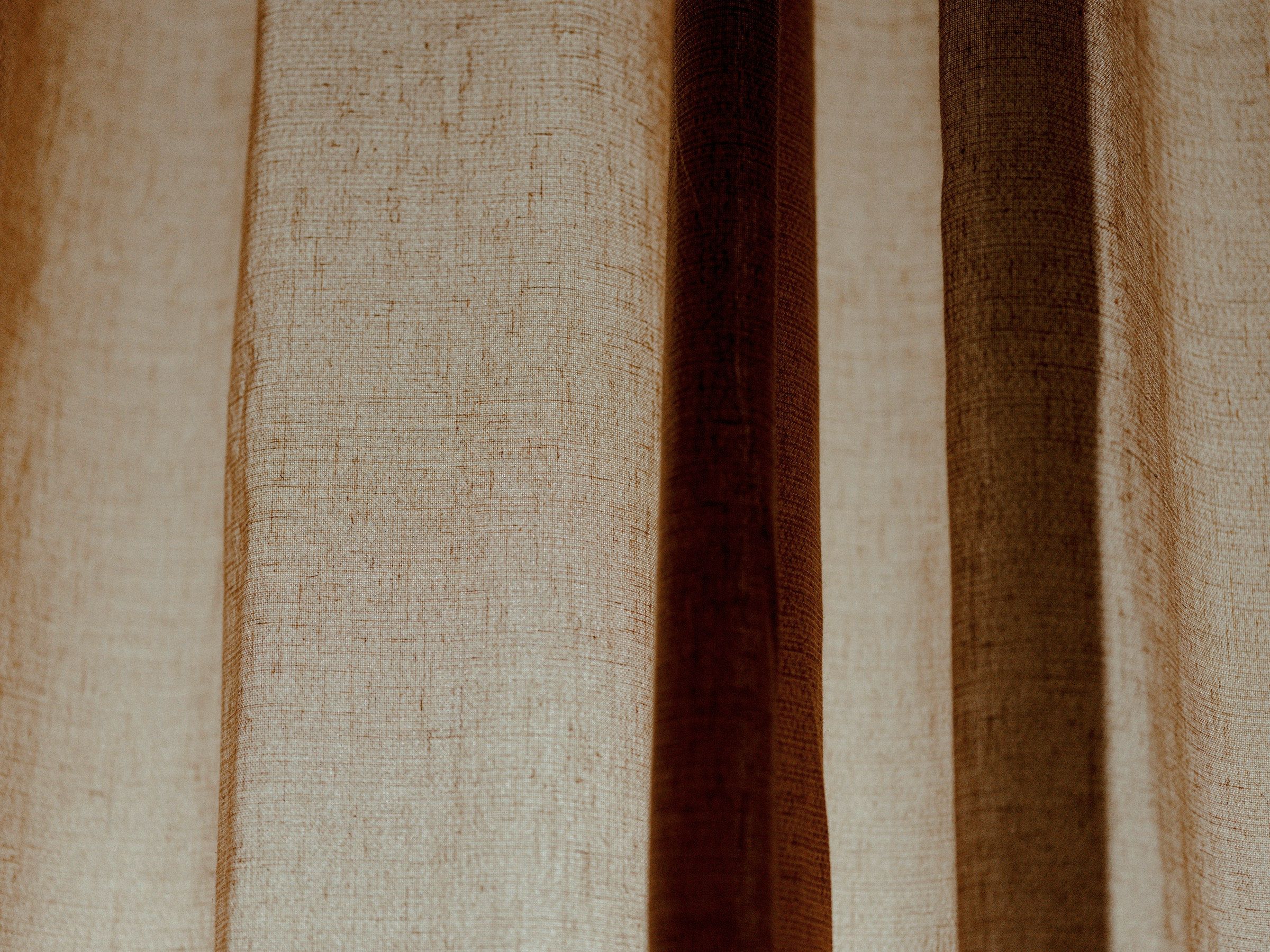 A linen curtain in motion: backlit undyed linen with a warm stripe of shadow running through the weave.