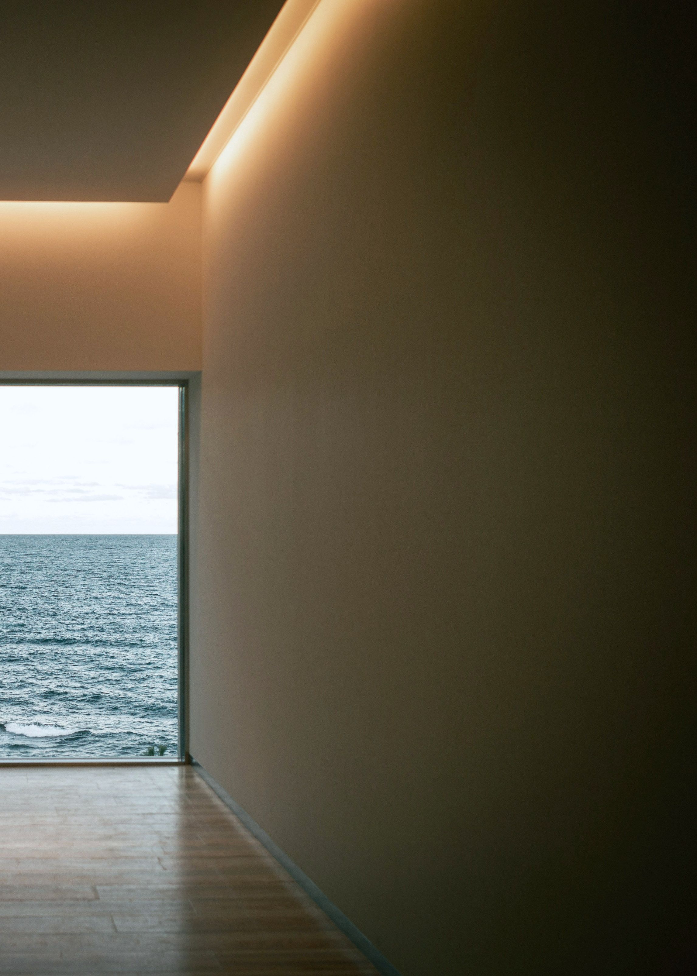 The living room toward the sea: a sand-plastered wall under a warm cove-lit ceiling, a honey timber floor, and a full-height opening onto flat open water.