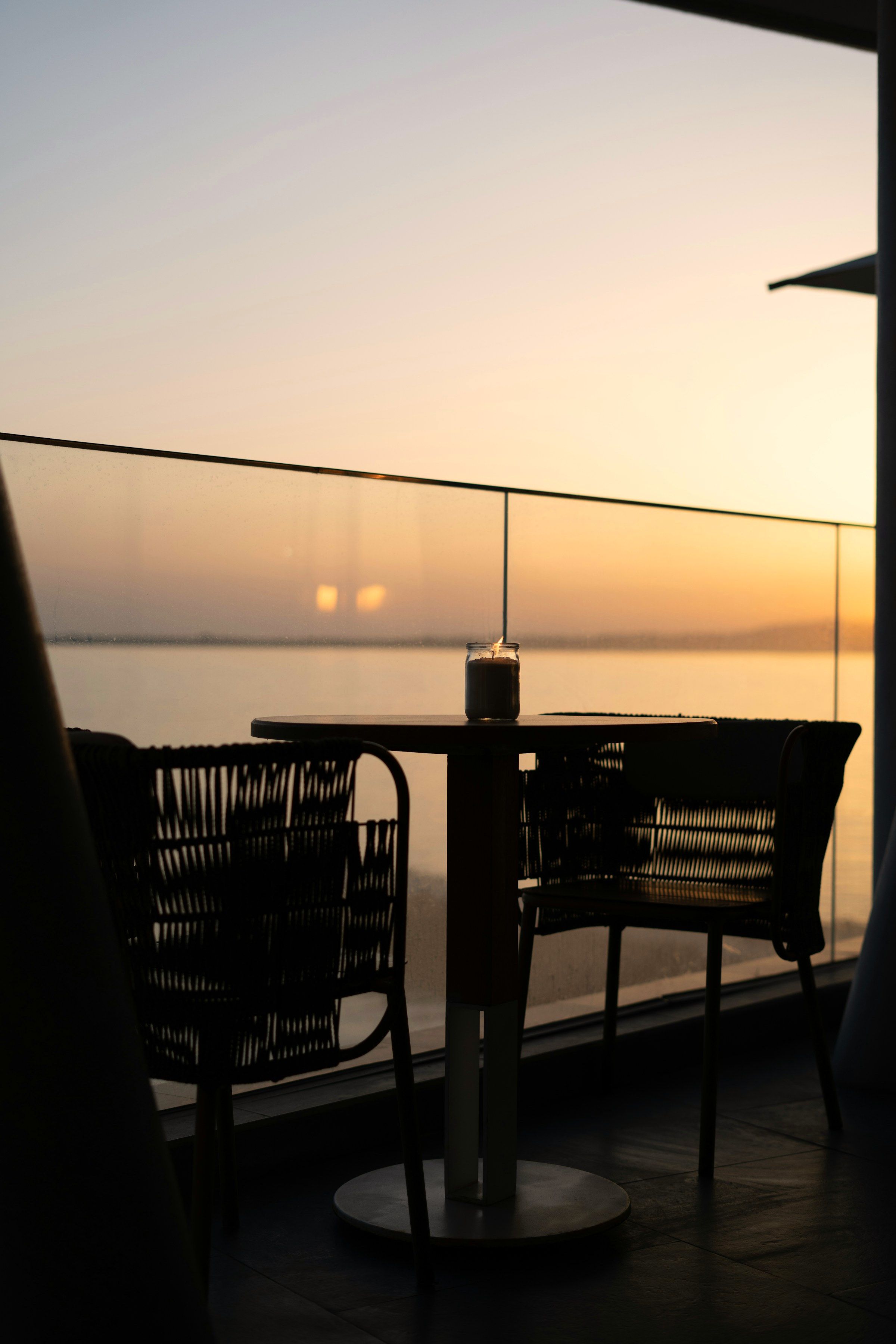 The terrace at dusk: a candle on a round table between two woven chairs, a glass balustrade, and flat calm water under a warm sky.