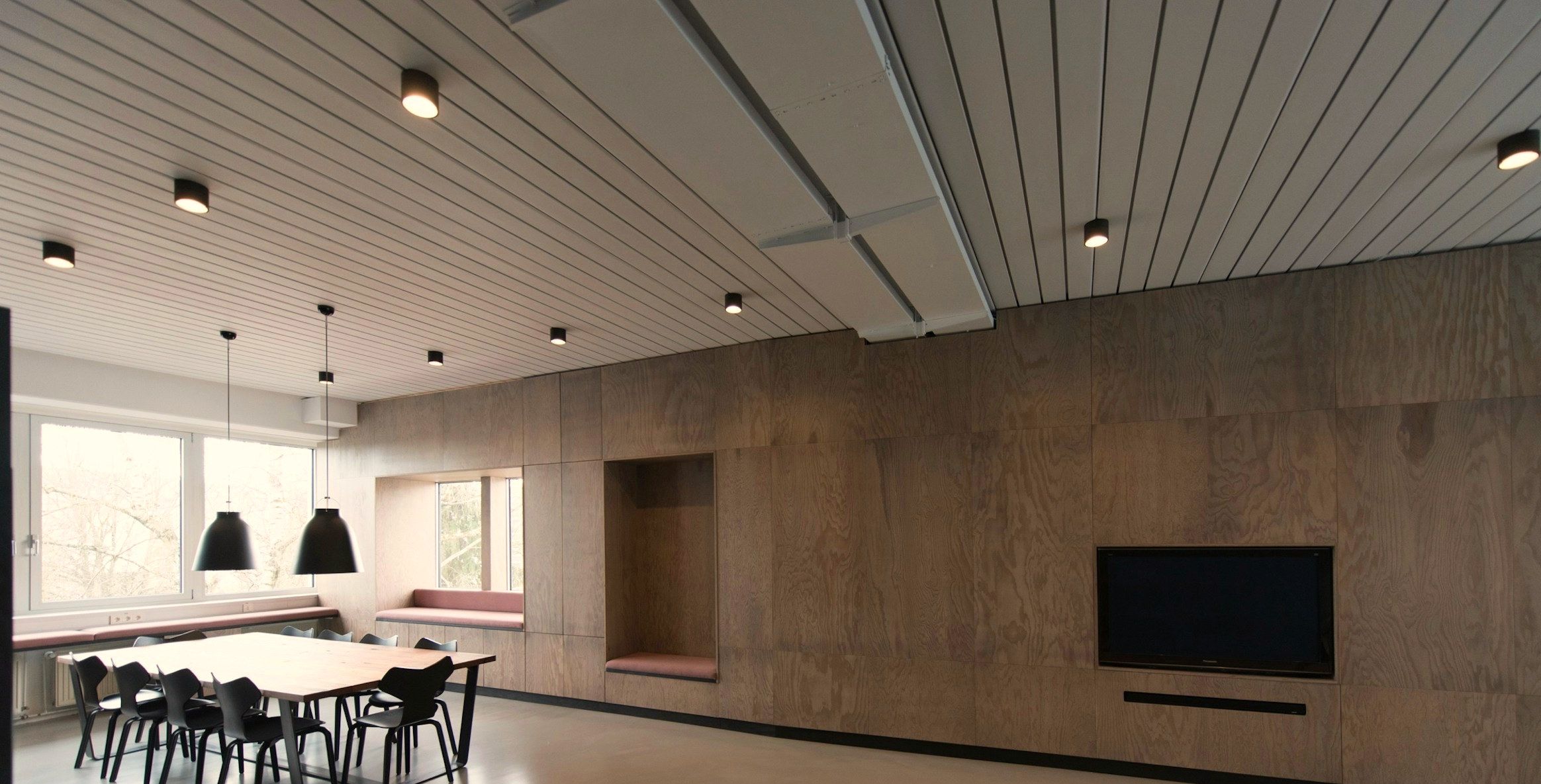The studio's meeting room: a long oak table and black chairs under low pendants, against a full-height plywood wall with a cushioned window seat.