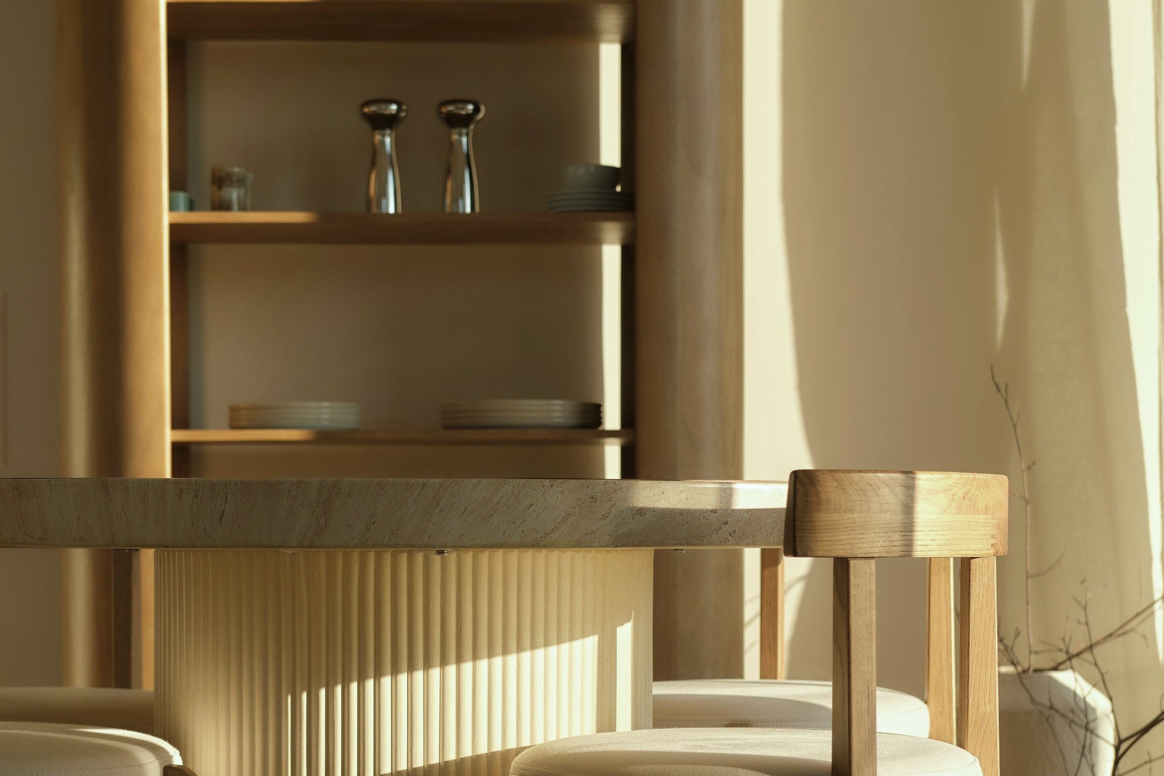 Late sun across the dining room at Apartment Falsledvej: oak chairs with cream cushions at a stone table on a fluted plaster base, ceramics on open oak shelving behind.