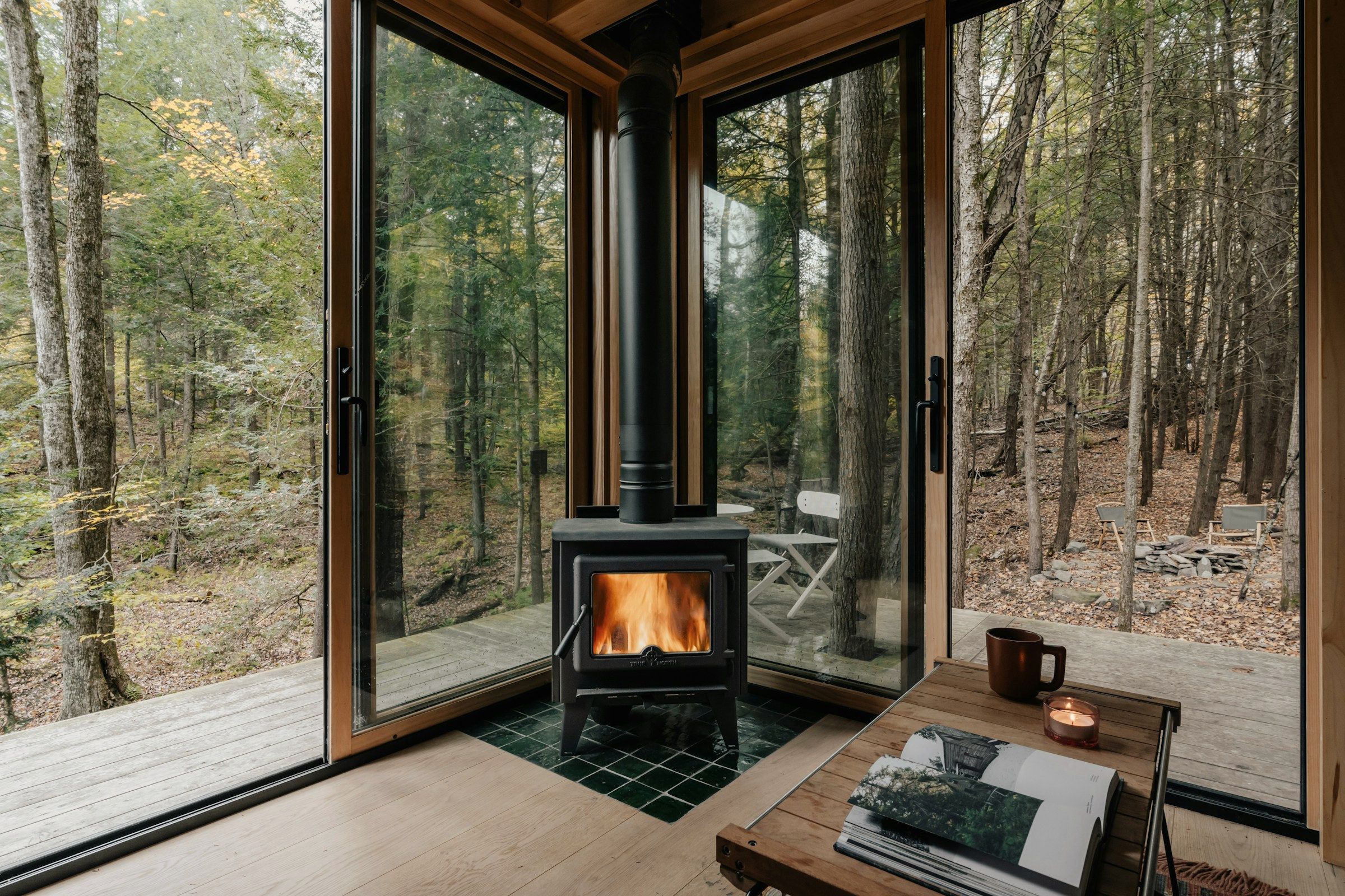 A room the studio built: the fire lit in a wood-burning stove set into a glazed corner, an oak table with an open book, a candle and a stoneware cup beside it, and birch woodland close on both sides of the glass.