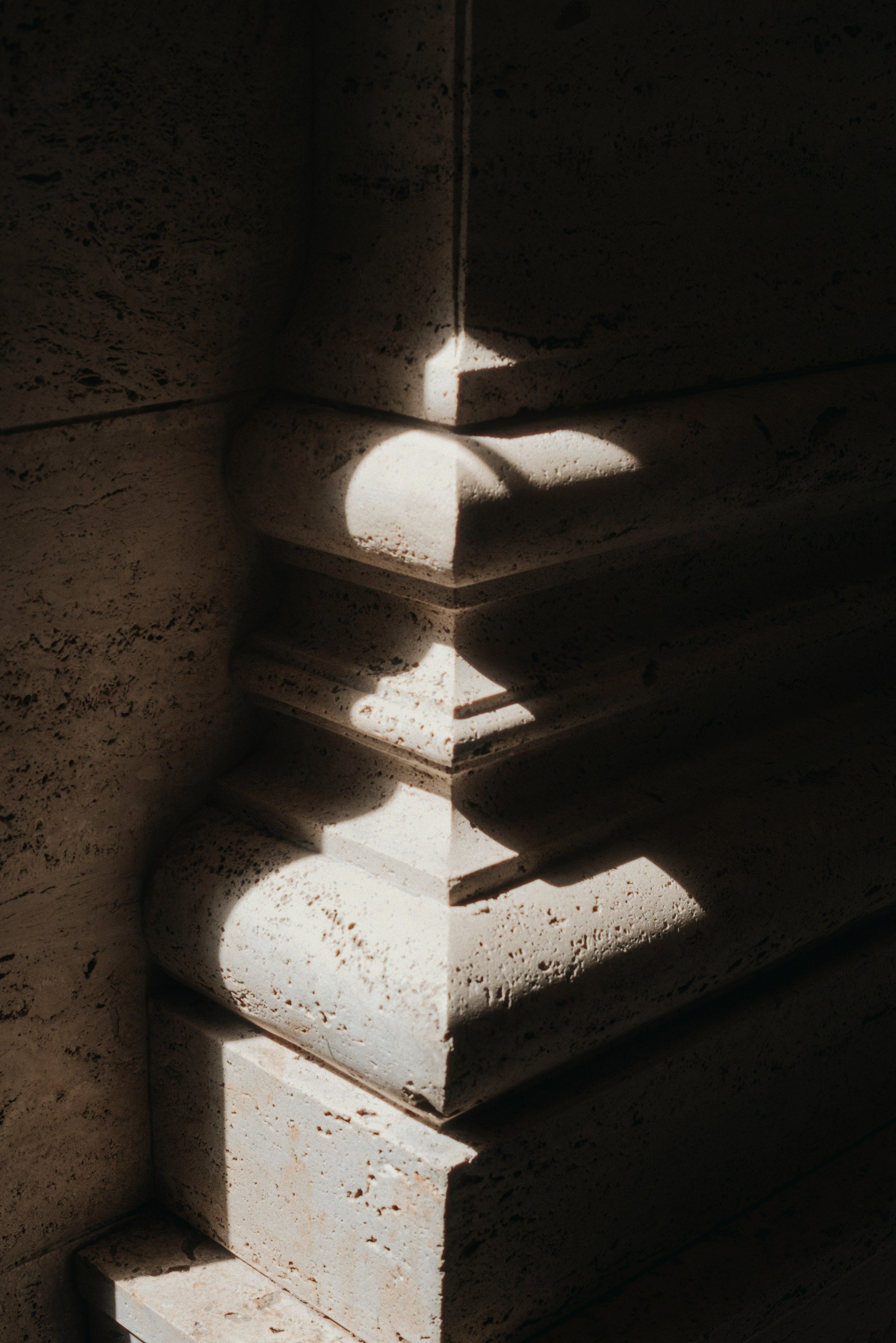 A travertine junction in the lounge at Hotel Søgrund: the moulded stone base of a pier stepping out from the wall, its pitted surface caught by a narrow blade of afternoon sun.