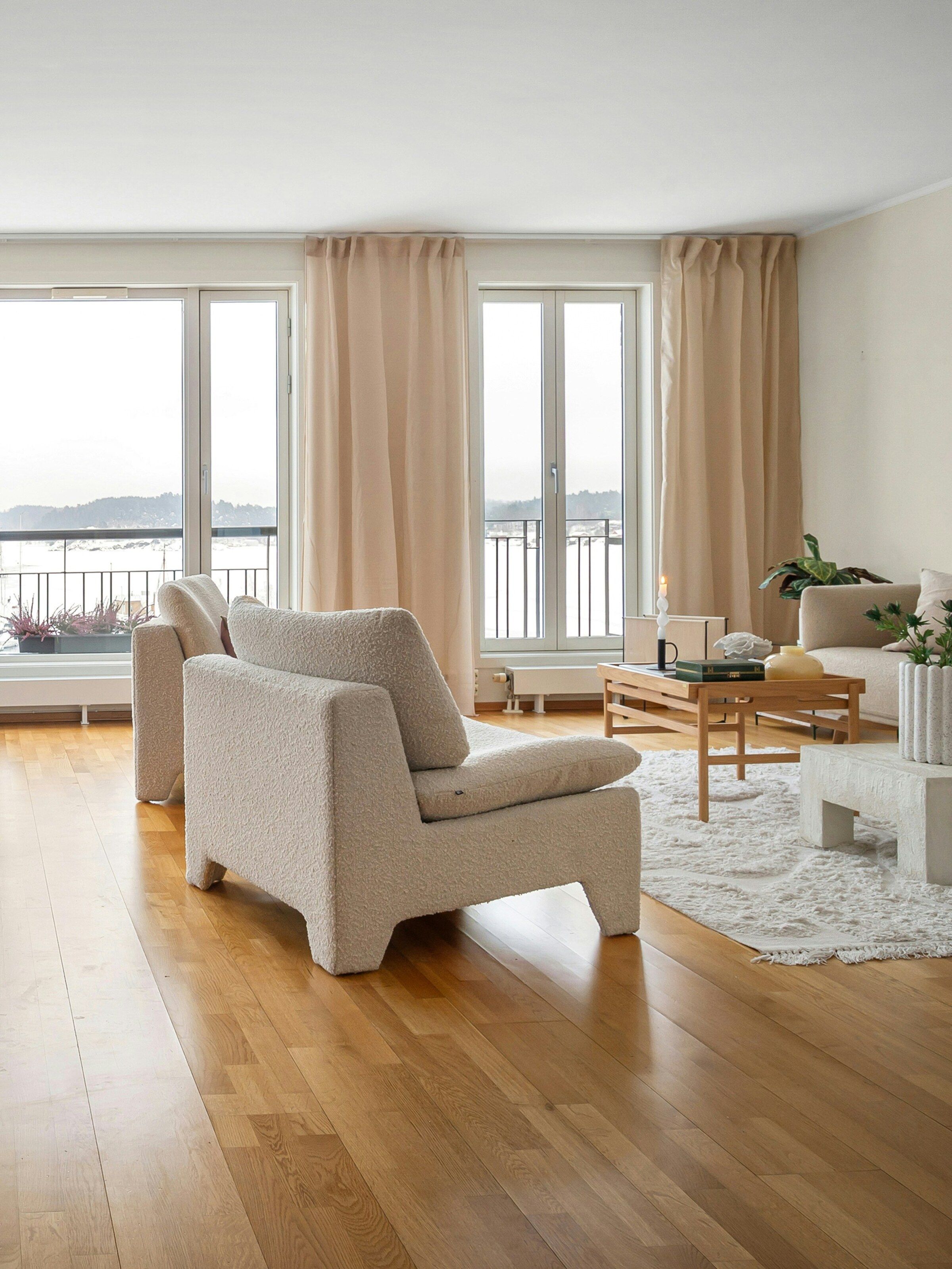 The living room at Villa Strandvejen: floor-length undyed linen curtains at the terrace doors, a bouclé armchair and an oak coffee table on a pale oak floor, with the flat water of the Øresund beyond.