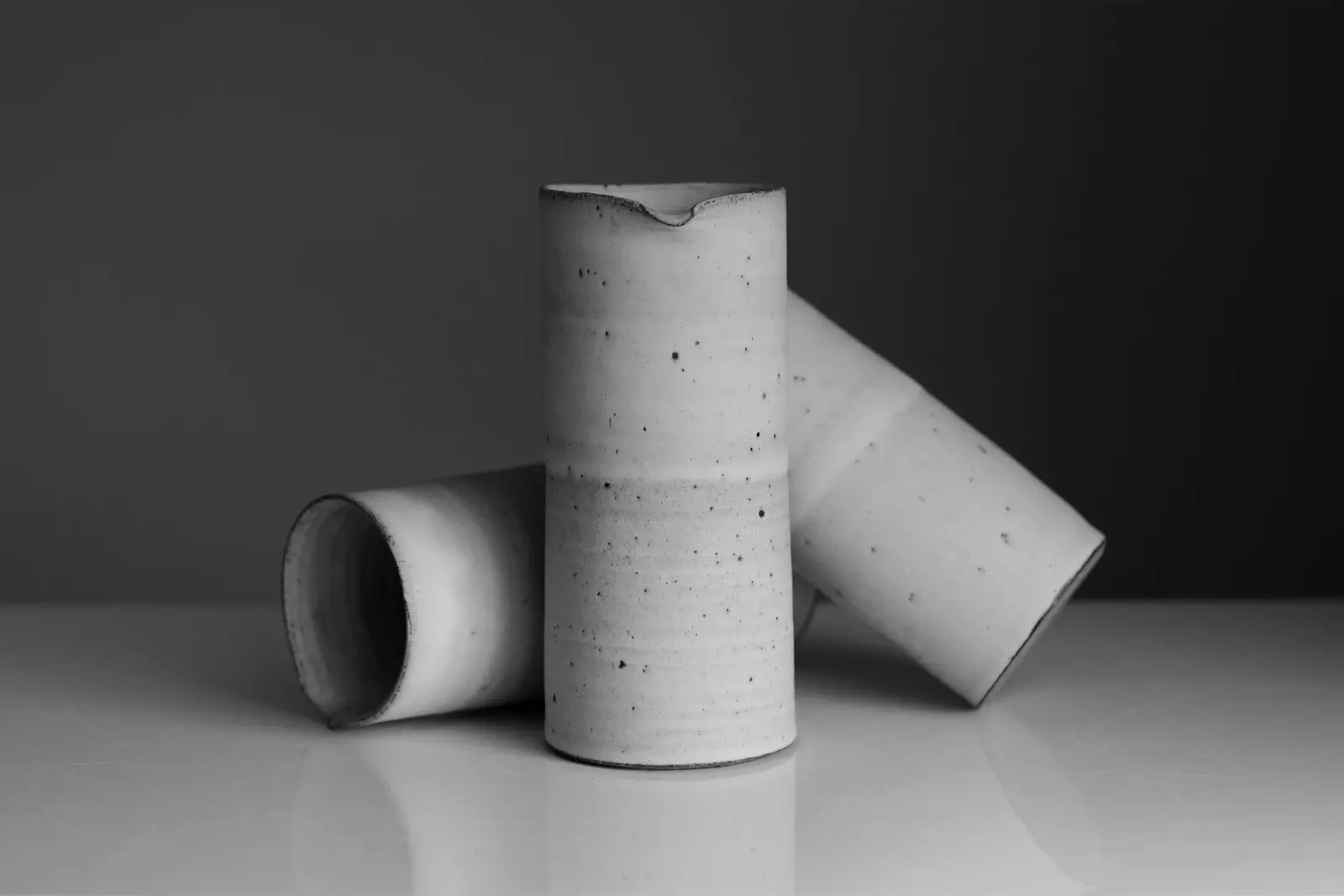 Speckled stoneware cups, one lying on its side, against a dark wall