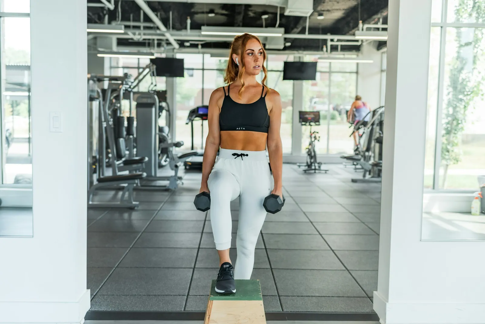 A woman walking across a bright gym floor carrying two dumbbells